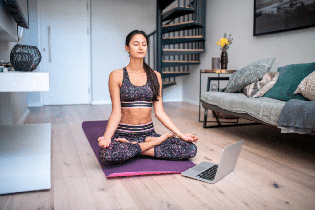 A woman meditating on a yoga mat in front of a laptop.