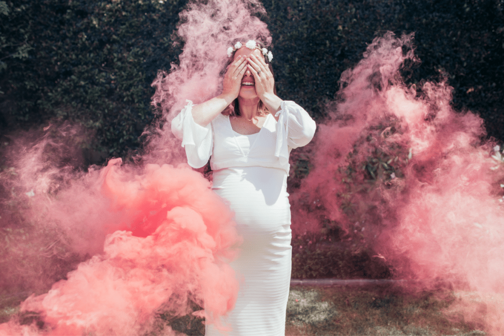 A pregnant woman in a white dress is covered in pink smoke.