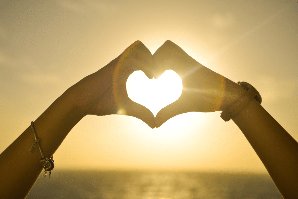 A woman's hands make a heart shape with the sun behind them.