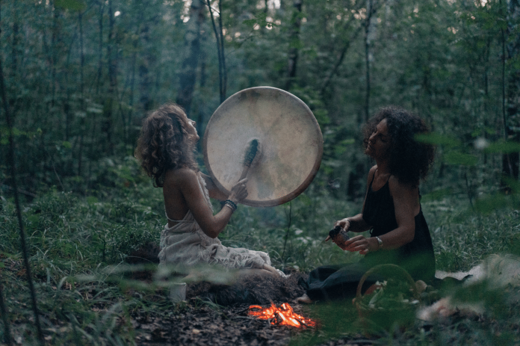 Two women playing a drum in the woods.