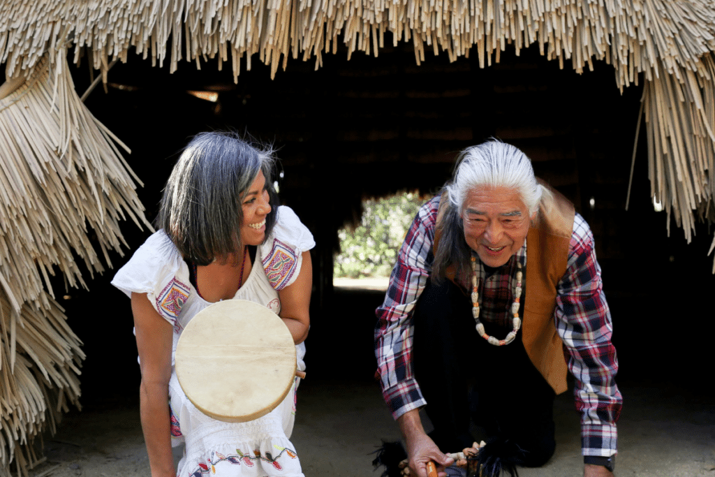 A man and woman playing a drum in front of a hut.